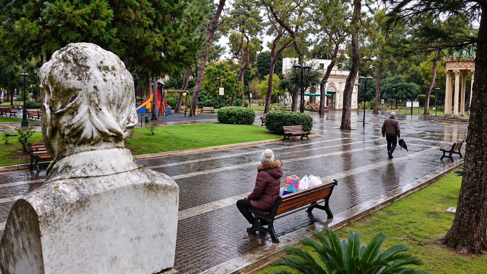 Giardino pubblico dopo la pioggia, una donna seduta su una panchina osservata da un busto in marmo